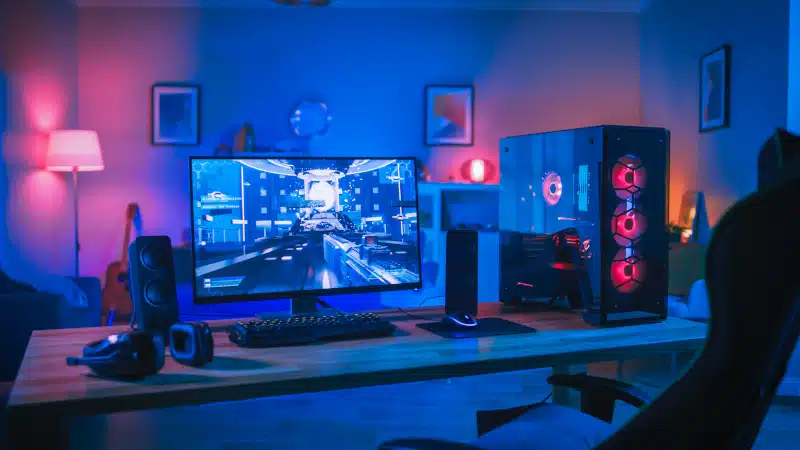 Powerful Personal Computer Gamer Rig with First-Person Shooter Game on Screen. Monitor Stands on the Table at Home. Cozy Room with Modern Design is Lit with Pink Neon Light.